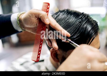 Crop anonymous barber using scissors and comb to cut hair of male client while working in grooming salon Stock Photo