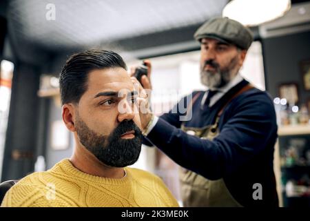 Concentrated middle aged bearded ethnic male barber in apron and beret spraying styling product on dark hair of serious customer in beauty salon Stock Photo