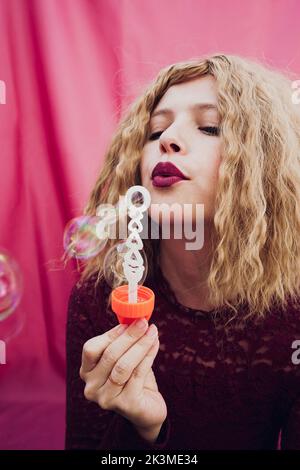 Stylish young female with blond wavy hair blowing soap bubbles on pink background in studio Stock Photo