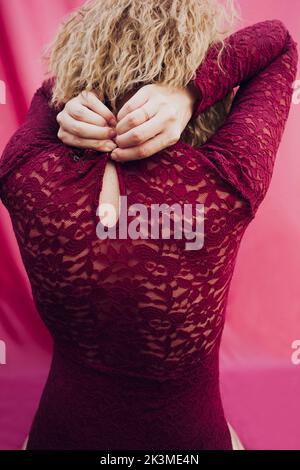 Back view of anonymous female with blond hair buttoning red dress on pink background in studio Stock Photo