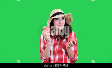 funny, smiling female farmer in checkered shirt and hat, holding in hands two large sour apples, eats, bites them, wrinkles her face, on green background in studio. Healthy food to your table. High quality photo Stock Photo