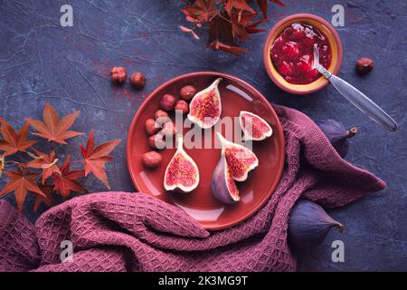 Autumntime background with fresh halved fig fruits on terracotta ceramic plate. Magenta towel and red maple Autumn leaves on dark purple board. Fig Stock Photo