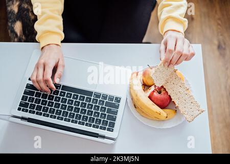 Freelancer young woman eating healthy food when working from home. Woman eating Healthy Grain Snacks and fruits while working with laptop at home Stock Photo
