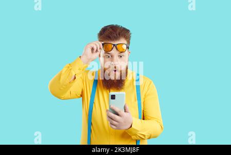 Amazed charismatic chubby man looks in surprise at screen of mobile phone with raised glasses. Stock Photo
