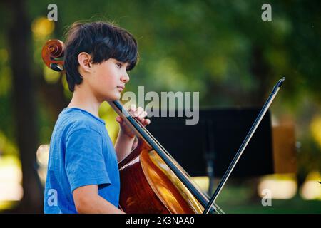 A serious boy sits in sunlit park playing cello Stock Photo