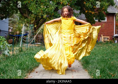 A girl in gold dress and wings leaps barefoot into air in backyard Stock Photo