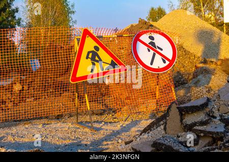 road signs indicating repair work and prohibiting passage are installed on a temporary fence with an orange emergency grid, selective focus Stock Photo