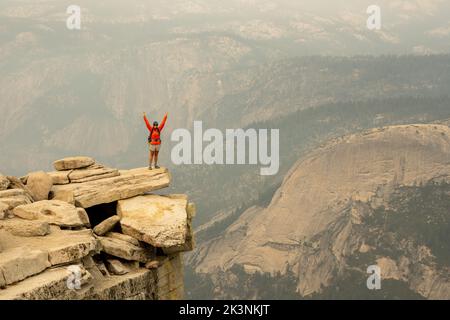 Woman In Orange Coat Stands With Arms Raised On Half Dome Ledge on smoky afternoon Stock Photo