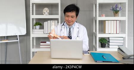 Asian Orthopedic Surgeon in white gown and stethoscope pointing at human skeleton foot ankle bone joint anatomy model, present to patient via wireless Stock Photo