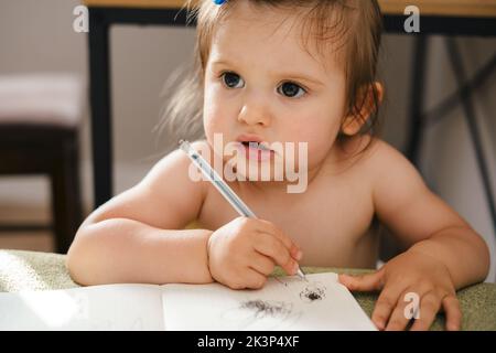 Portrait of a little caucasian girl playing, drawing in a textbook sitting at table in the living room. Toys for preschool, kindergarten. Children at Stock Photo