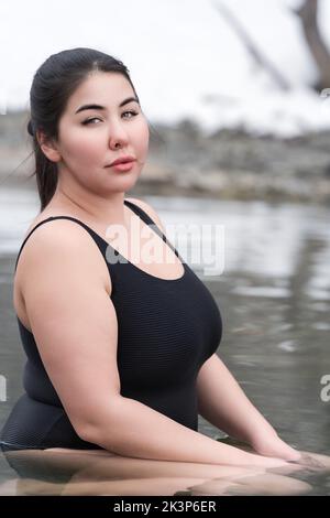 Young busty curvy full figured young adult model in black one piece bathing swimsuit sitting and relaxation in mineral water in outdoors pool at spa Stock Photo