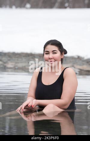 Busty curvy plus size young adult model in black one-piece bathing suit sitting in geothermal water in outdoors pool at balneotherapy health spa Stock Photo