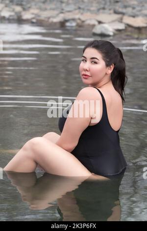 Overweight young woman in black one-piece bathing swimsuit sitting in mineral water in outdoors pool at balneotherapy spa, hot springs resort Stock Photo