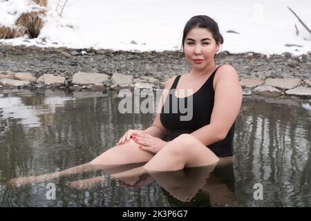 Outsize young adult woman in black one piece swimming costume sitting bathing in mineral water in outdoors pool at balneotherapy health spa Stock Photo