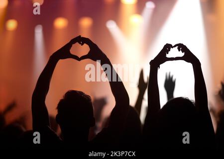 Music lovers. Rearview of audience members at a music concert holding up their hands in a heart symbol. Stock Photo