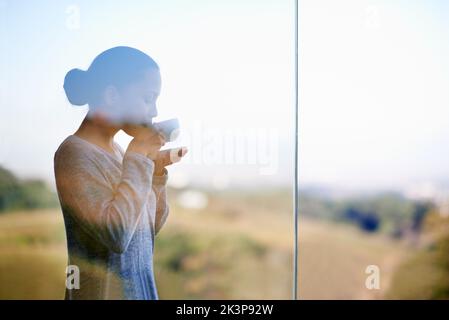 Enjoying a warm cup of coffee while surrounded by nature. Thorugh the window shot of a young woman drinking coffee while standing outdoors. Stock Photo