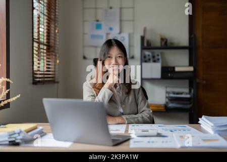 Portrait pretty asian businesswoman smiles at the camera while sitting at her desk in front of the laptop computer Stock Photo
