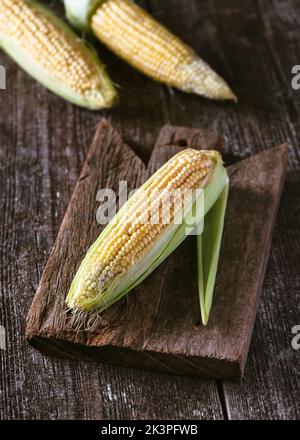 Fresh raw sweet corn on cobs with husk on rustic wooden cutting board  ready for cooking. Rustic style. Healthy vegan food concept. Stock Photo