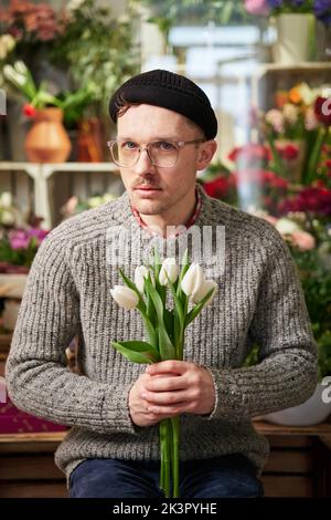Mother's Day, dating, love, romantic or International Women's Day concept. Attractive bearded florist hipster caucasian male in sweater, eyeglasses and knit hat with bouquet of white tulips Stock Photo