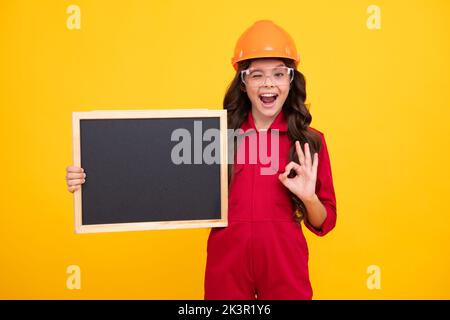 Surprised teenager girl. Child girl builder in hard hat helmet. Teenage girl worker hold blackboard isolated on yellow background. Kids renovation Stock Photo