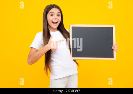 Teenager younf school girl holding school empty blackboard isolated on yellow background. Portrait of a teen female student. Excited teenager, glad Stock Photo