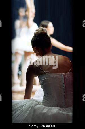 A ballerina backstage of a performance at a theatre on stage. Ballet dancer sitting on the floor while play or concert is happening. She is getting Stock Photo