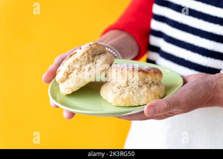 A smartly dressed woman is shown holding two freshly made traditional plain scones on a small plate. The cakes are lightly dusted with icing sugar Stock Photo