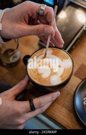 Hot cappuccino in a cup, and a barista making a bear face shape on top, using a wooden stick. Stock Photo