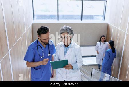 Mature doctor and male nurse reviewing patient's medical chart standing on stairs in hospital. Healthcare workers Stock Photo