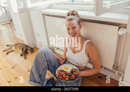 Blonde woman in top holding plate with fried egg and fresh croissant near blurred scottish fold cat at home,stock image Stock Photo