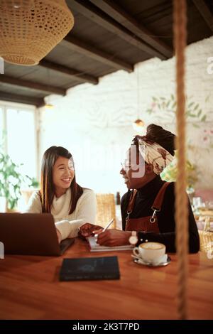 Two beautiful multicultural female students sitting in a cafe and following online lecture on the laptop. Technology in education. Stock Photo