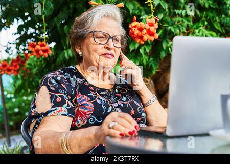 Elderly lady in casual outfit and eyeglasses sitting at table while using netbook and talking on smartphone near green plants and flowers in backyard Stock Photo