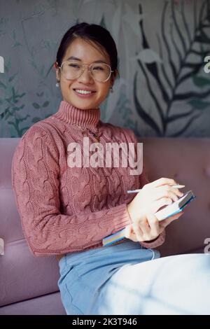 A happy female student is sitting in a coffee shop and writing down to do list in a textbook. Stock Photo