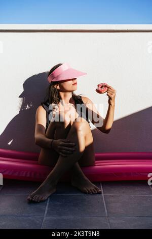 Full body of young female in swimsuit and hat sitting on pink inflatable mattress and enjoying delicious donut while chilling on poolside in summer da Stock Photo