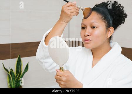 Delighted charming female in bathrobe applying foundation with brush on face while looking in mirror and doing makeup at home Stock Photo