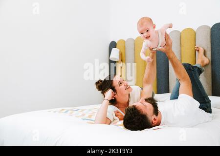 Father pretending that playful baby flying like superhero while chilling together with wife and kid on soft bed at home Stock Photo