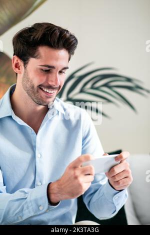 Happy handsome bearded male in casual blue shirt using mobile phone while sitting against light wall and green plants Stock Photo