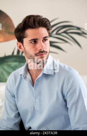 Modern handsome bearded male in casual blue shirt looking away while sitting against light wall and green plants Stock Photo