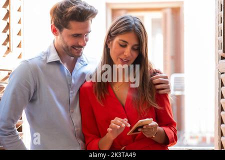 Positive young stylish couple using mobile phone and reading message while standing together against window at home Stock Photo