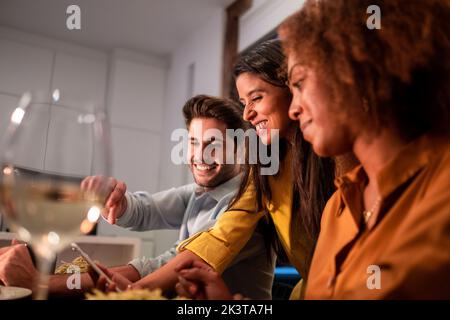Cheerful young multiracial friends using mobile phone while having dinner during home party Stock Photo
