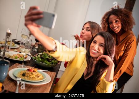 Cheerful young female friends taking selfie with smartphone while having dinner during home party Stock Photo