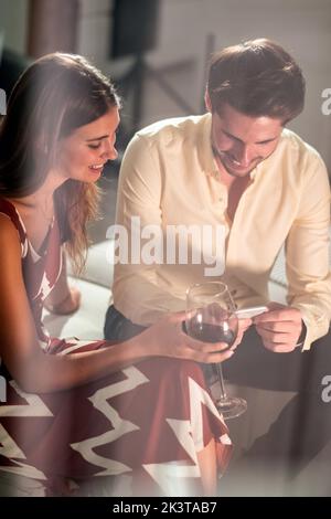 Throughout windows view of cheerful young man share photos on cellphone with female friend while resting together on sofa at home Stock Photo