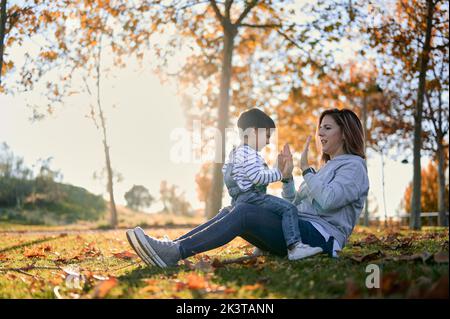 Ground level of loving mother clapping hands with little boy while having fun and playing in autumn park Stock Photo