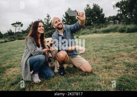 Lovely couple hugging Labrador Retriever and having fun taking selfie on mobile phone in cloudy day in park Stock Photo