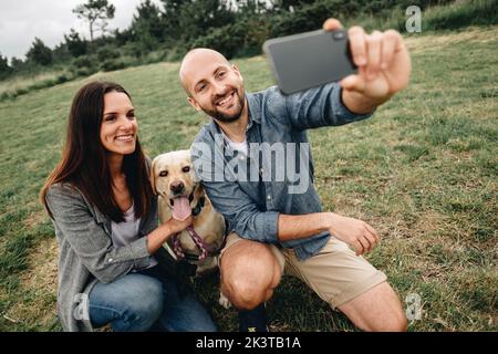 Lovely couple hugging Labrador Retriever and having fun taking selfie on mobile phone in cloudy day in park Stock Photo