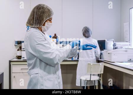 Young woman in glasses standing in modern laboratory and putting on gloves. Stock Photo