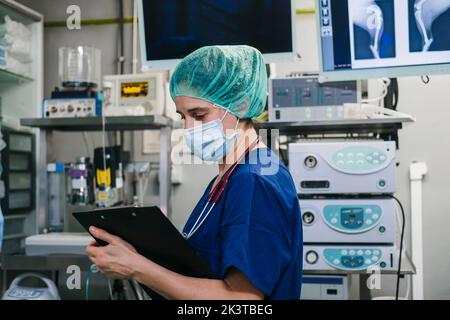Side view of young female veterinary in medical mask and uniform standing in operating room and taking notes in clipboard with pen before surgery in c Stock Photo