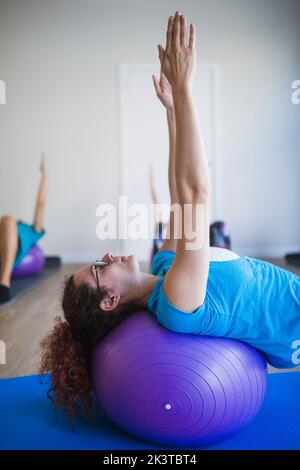 Side view of young female fitness instructor lying on fit ball with arms outstretched while doing exercises with group of students in sports studio Stock Photo
