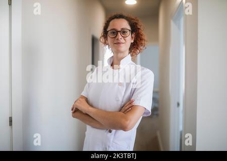 Positive young curly haired female physiotherapist in white uniform and eyeglasses smiling friendly and looking at camera while standing in hallway of Stock Photo