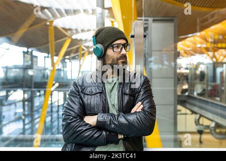 Handsome bearded hipster guy in eyeglasses and headphones wearing black leather jacket and knitted hat looking away while standing in airport hall Stock Photo